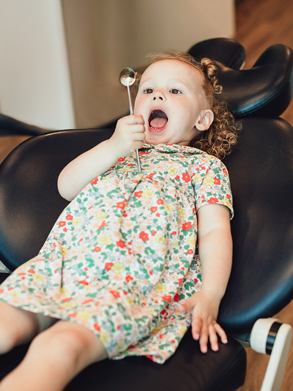 Young child in dental chair looking into her mouth with a mirror