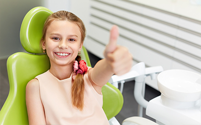 A young girl sitting in a dental chair giving a thumbs up