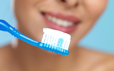 A close-up of a toothbrush with toothpaste on it and a woman smiling in the backdrop